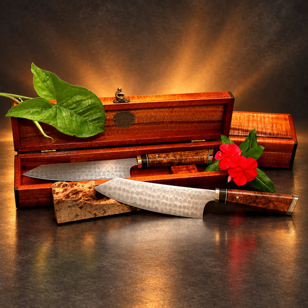 Japanese bunka style kitchen knives with whirlpool damascus hand-forged blades and koa wood decorative handles rest in front of an open matching wooden keepsake box. A second box is partially visible. These luxury cutlery items were handmade by SalterFineCutlery