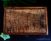 A rectangular wooden box with a richly figured old-growth Hawaiian curly koa wood grain pattern and a monogrammed 'H' in the bottom right corner, displayed on a dark background with decorative green leaves and a small orchid handcrafted by SalterFineCutlery