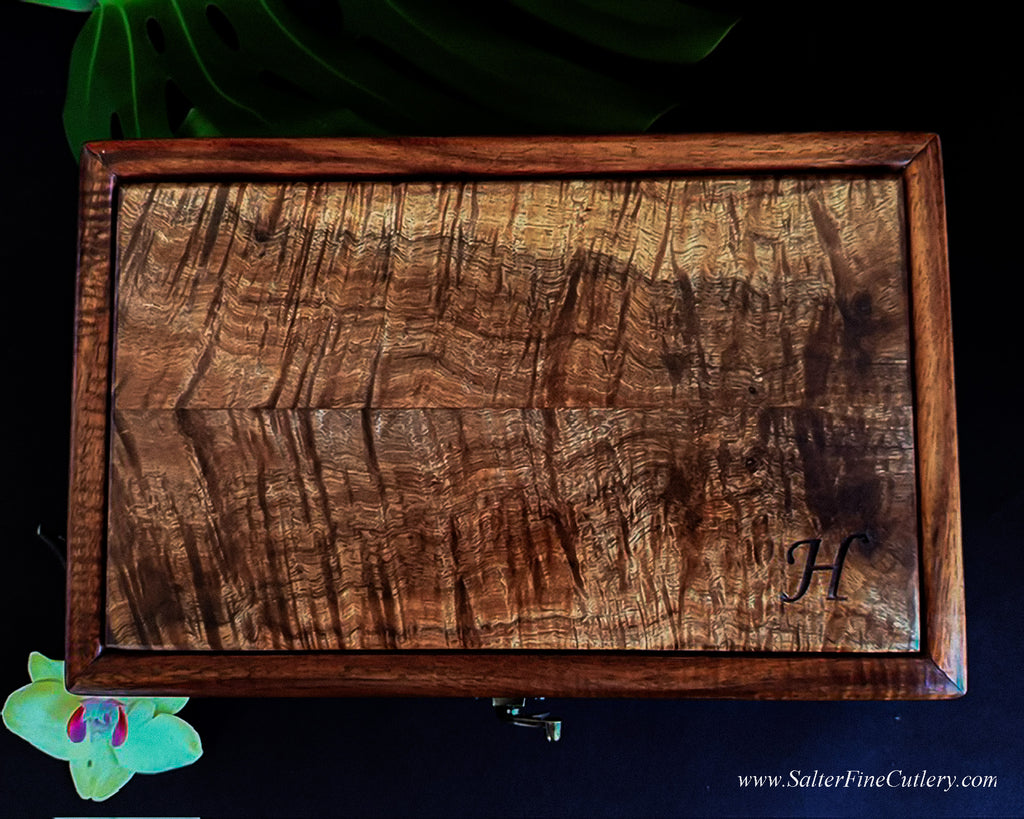 A rectangular wooden box with a richly figured old-growth Hawaiian curly koa wood grain pattern and a monogrammed 'H' in the bottom right corner, displayed on a dark background with decorative green leaves and a small orchid handcrafted by SalterFineCutlery